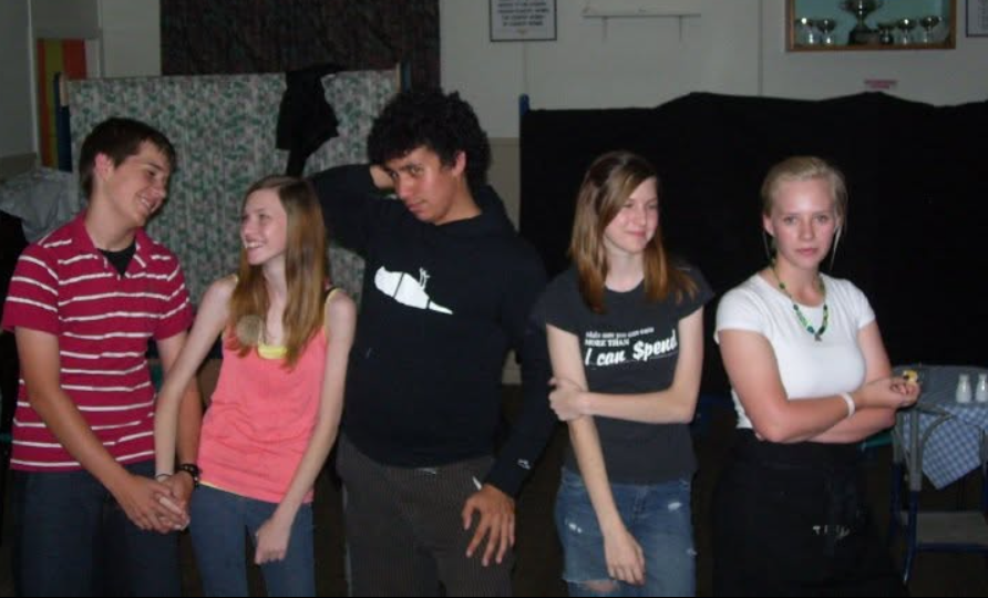 Five teenagers standing together; the girl on the right stands apart with arms crossed and a skeptical, jealous-looking expression